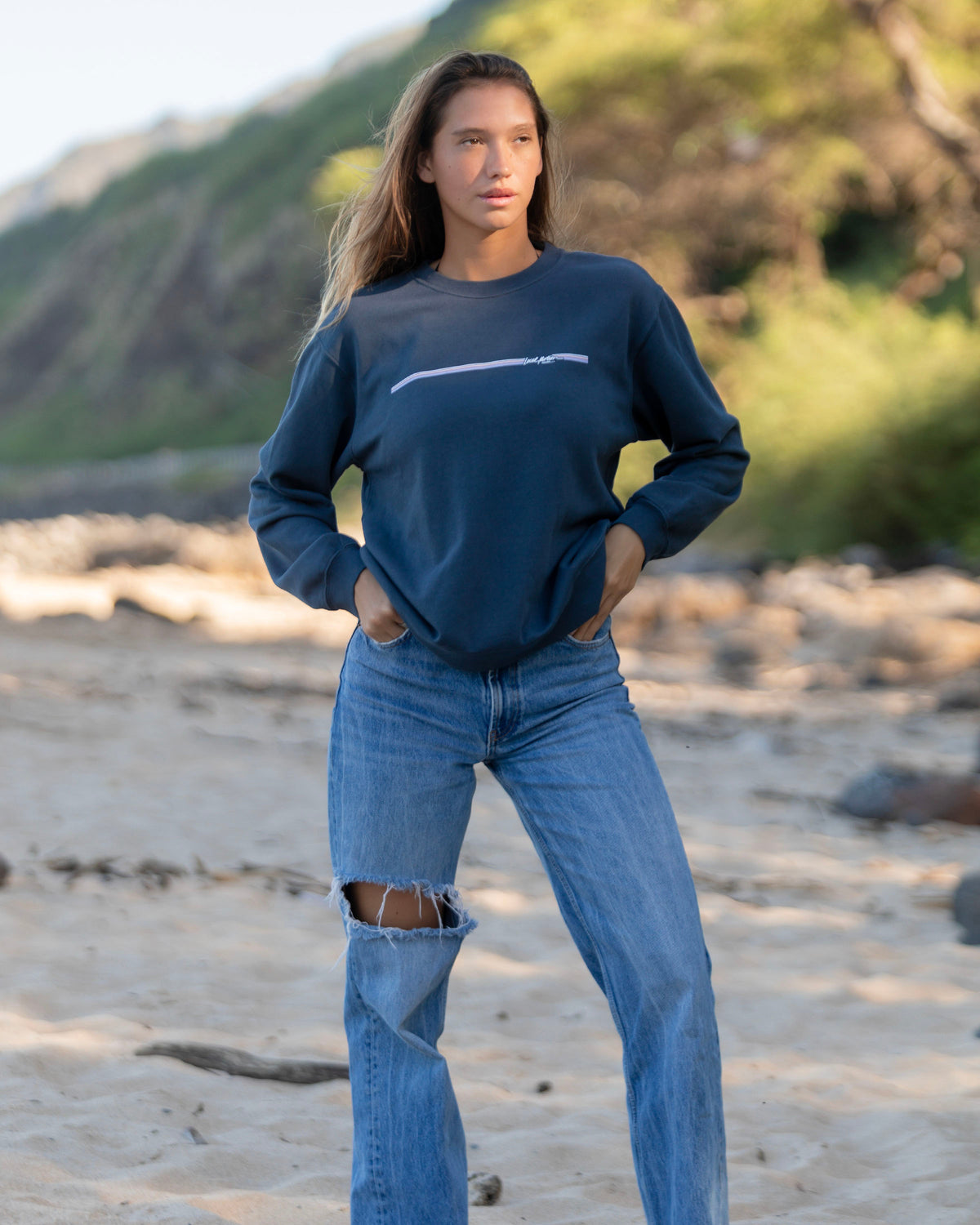 A woman standing on a sandy beach wearing a navy blue crewneck sweatshirt 