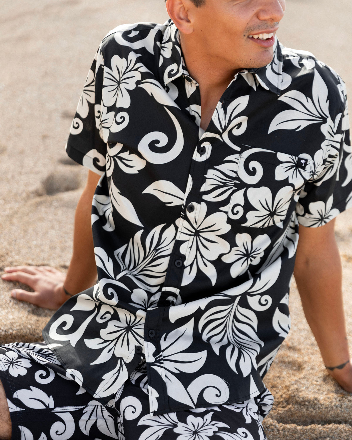 A man wearing a black short-sleeve button-up shirt and matching shorts with a bold white tropical floral and swirl pattern, sitting on a sandy beach. [black]