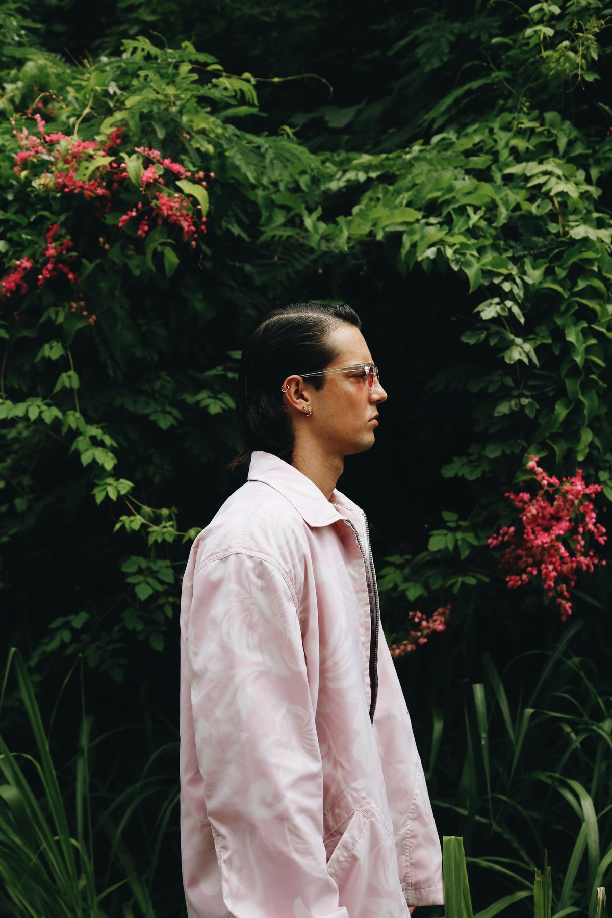 A man in profile wearing a light pink overshirt and glasses, standing in front of a lush green garden background with pink flowers.