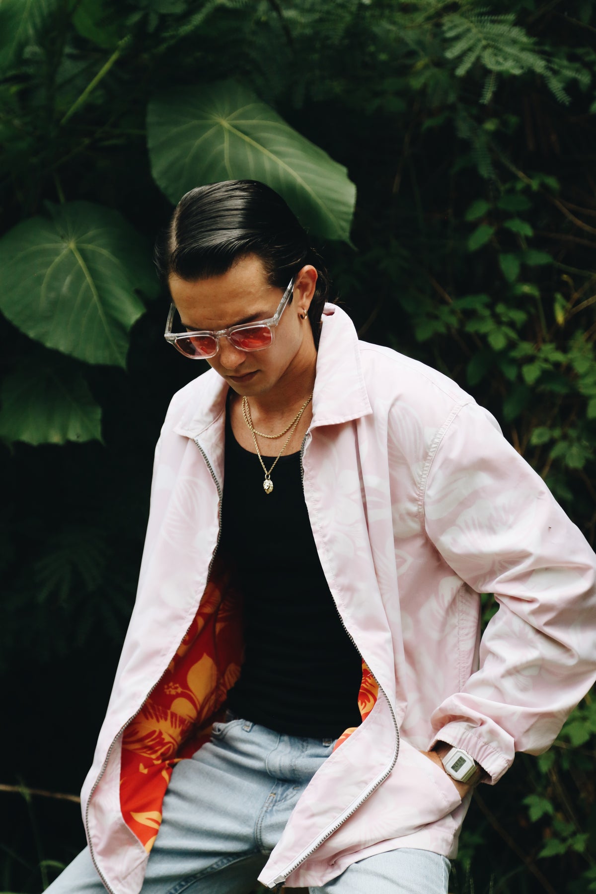 A man wearing a light pink overshirt over a black t-shirt, light blue jeans, and pink-tinted sunglasses, posed against a lush green foliage background.