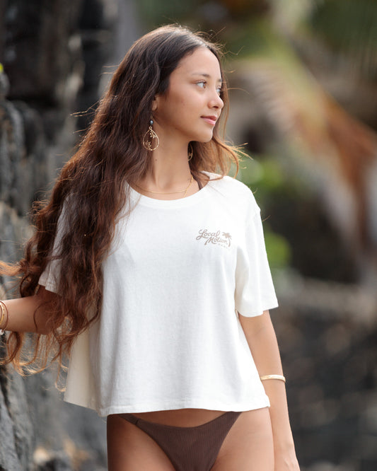 A woman with long, wavy brown hair wearing a white short-sleeve crop top with a small script logo on the left chest [cream]