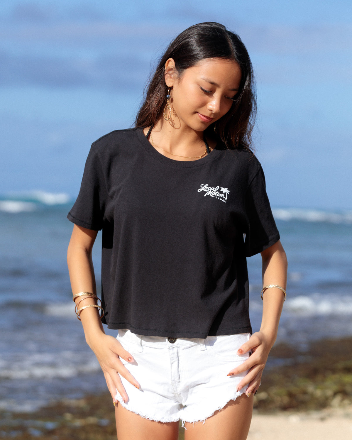 A woman standing on a beach wearing a black short-sleeve crop top with a small white script logo [black]