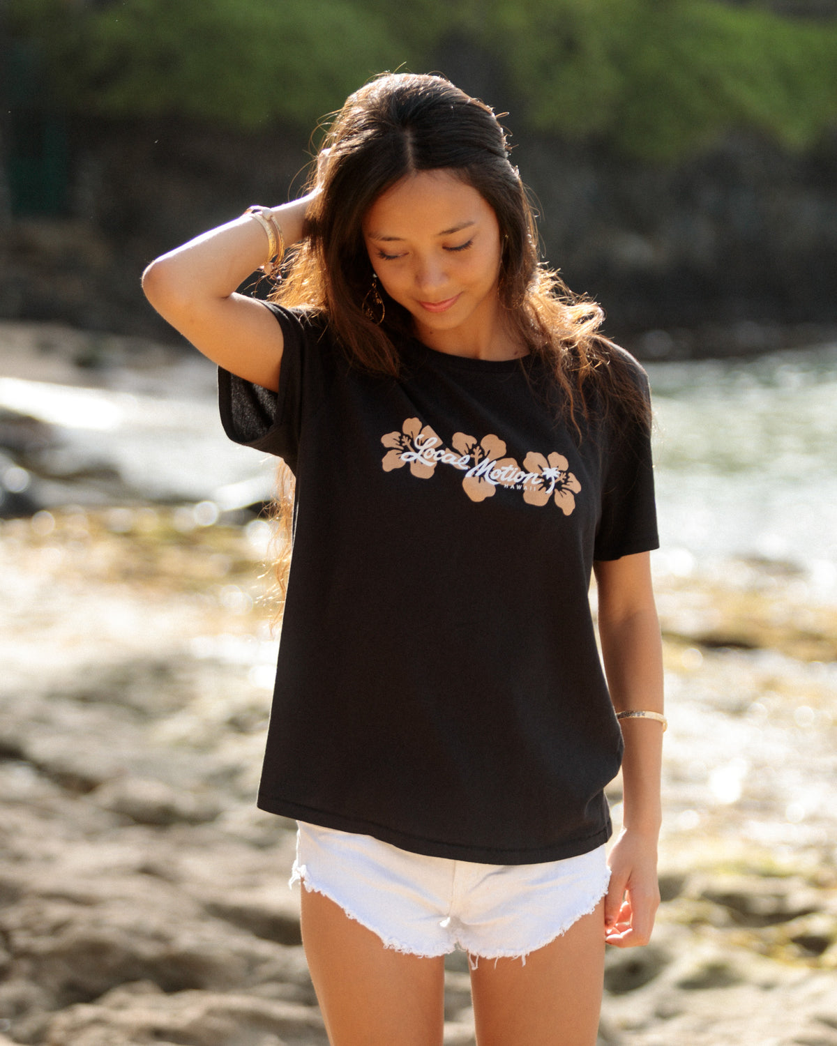 A woman standing on a beach wearing a black t-shirt with a white and tan hibiscus flower [black]