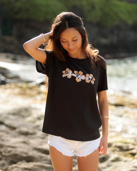 A woman standing on a beach wearing a black t-shirt with a white and tan hibiscus flower [black]